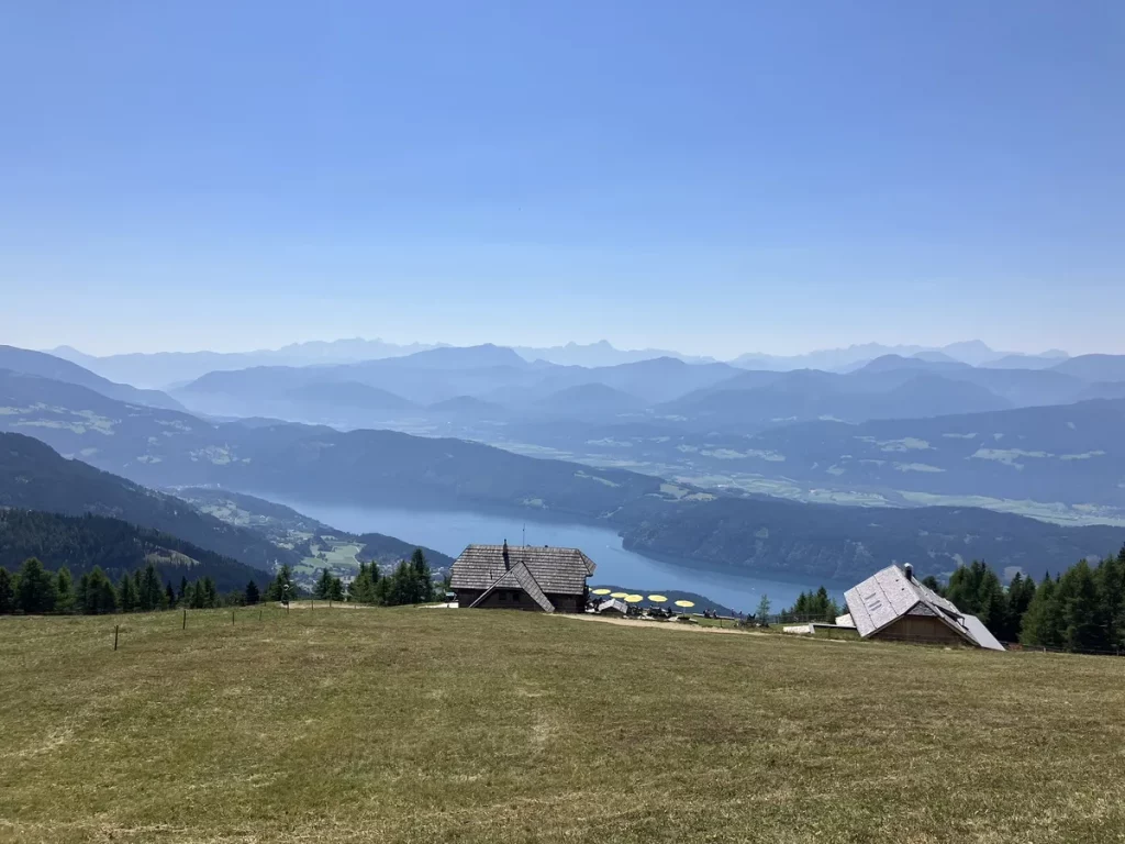 Wooden mountain huts on a grassy hill overlooking a blue alpine lake and layers of hazy mountains in Austria. A peaceful scene often found along a beginner hut to hut hike in Austria.