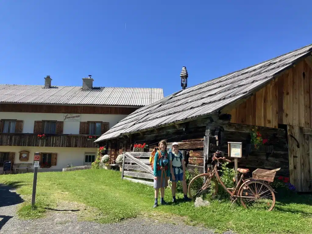 Two hikers with backpacks standing outside a rustic alpine hut with wooden shutters, bright flowers, and an old bicycle leaning nearby.