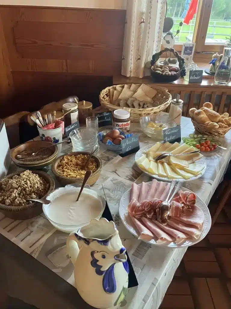 A breakfast spread of sliced meats, cheese, bread, cereals, yogurt, and condiments set out on a cozy wooden table inside a mountain hut.