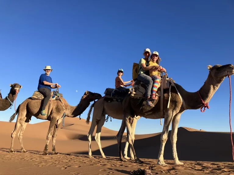 A family rides camels through golden sand dunes under a bright blue sky, with wide smiles and colorful clothing. Camel trekking in the desert offers adventurous October half term ideas for all ages.