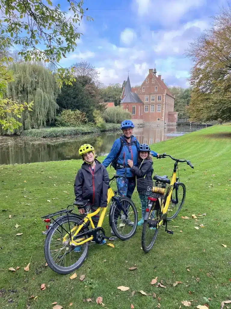 A smiling family wearing helmets poses with yellow bikes in front of a historic red-brick castle surrounded by a calm moat and autumn trees. Exploring European castles by bike is one of many scenic October half term ideas for families.