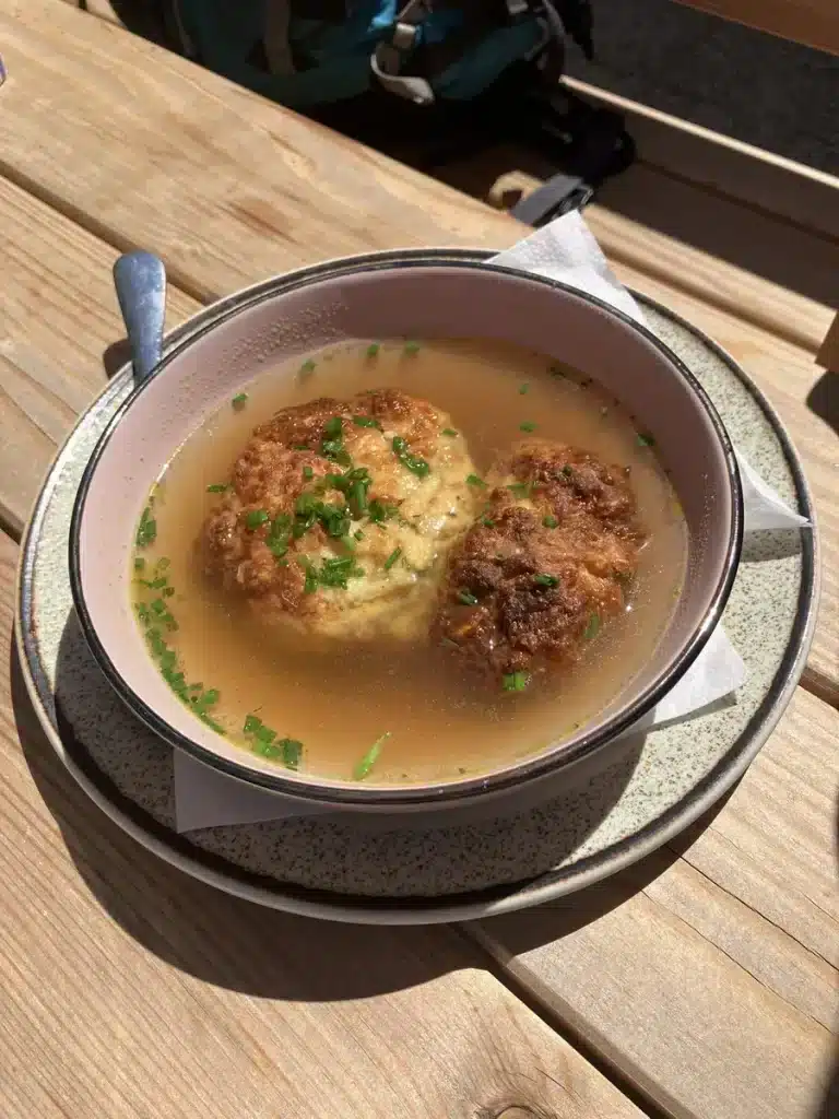A bowl of traditional Austrian soup with two dumplings garnished with chives, served on a rustic wooden table.