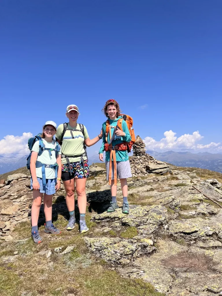 Three hikers smiling at the summit of a rocky mountain peak, with wide views of the Austrian Alps behind them.
