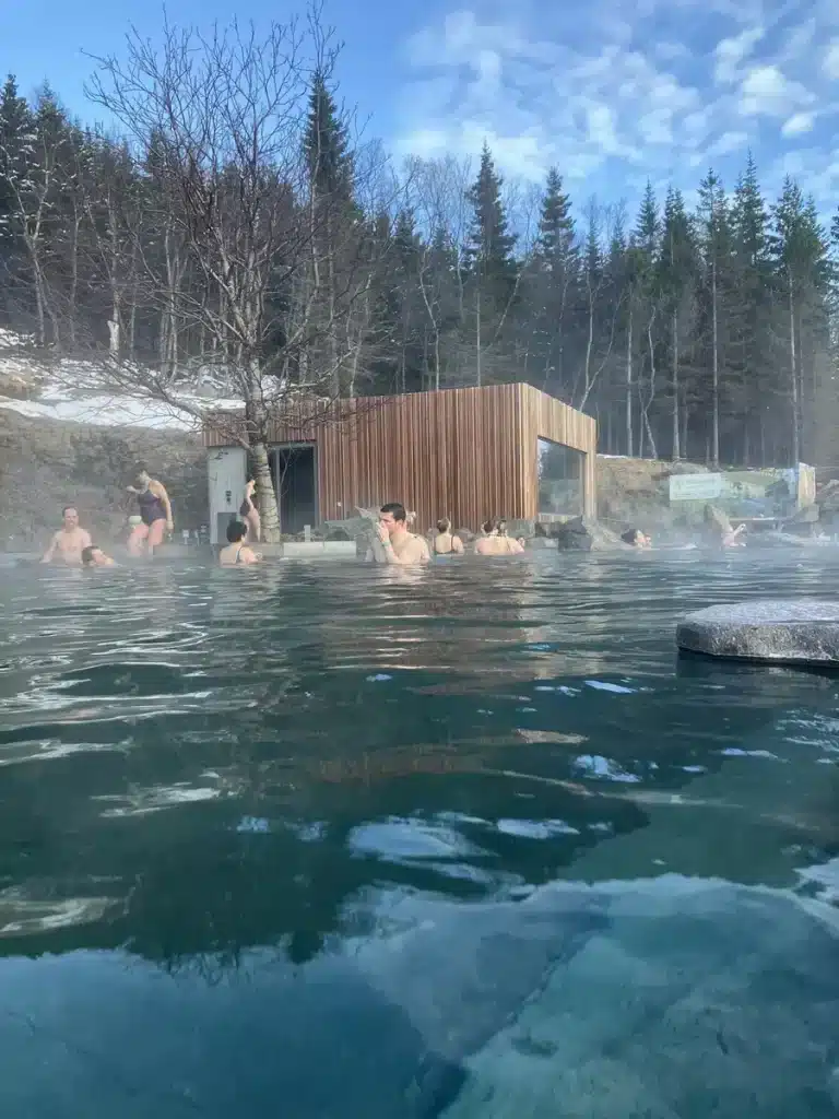 People relax in a steamy outdoor hot spring surrounded by snow-dusted trees and a modern wooden spa building. Soaking in natural hot springs is a relaxing October half term idea for chilly destinations.