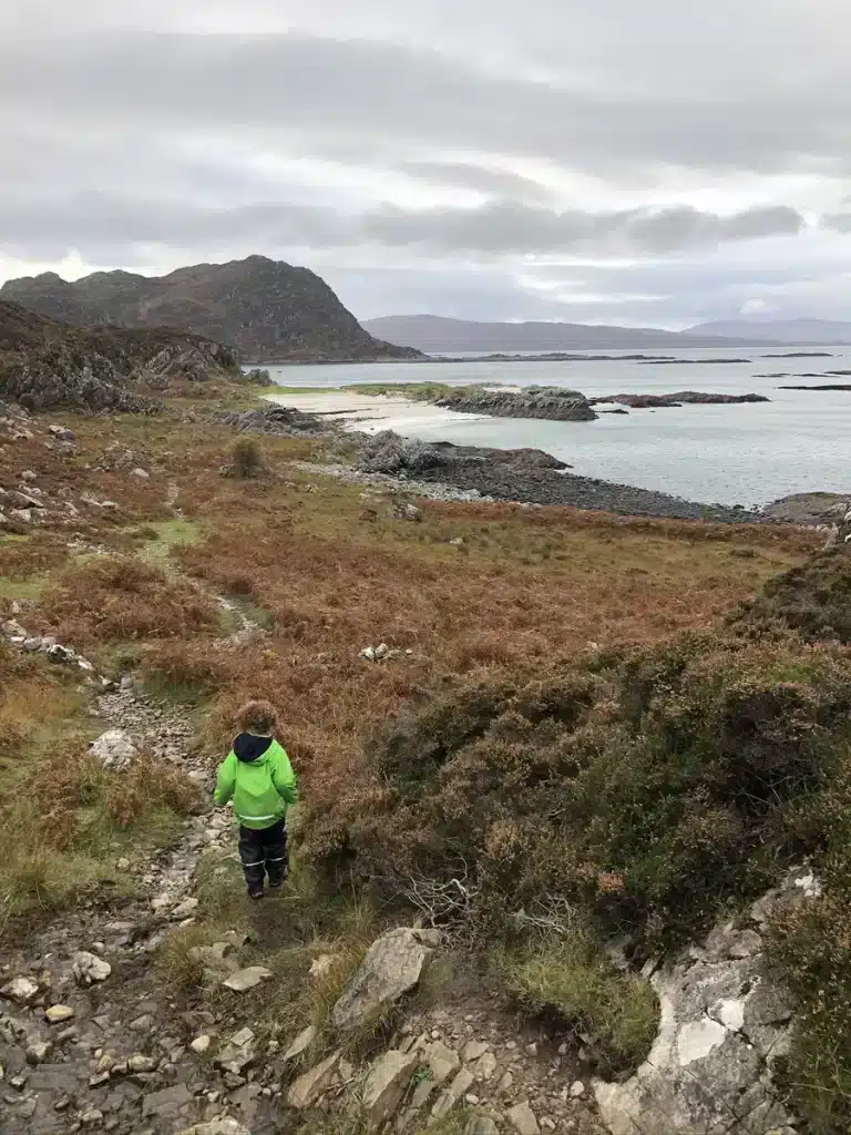 A child in a bright green jacket hikes along a rocky path near the Scottish west coast, with dramatic hills and calm waters in the distance. Coastal hikes offer active October half term ideas full of nature and fresh air.