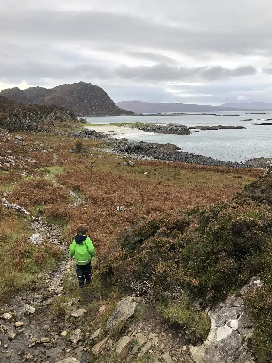 A child in a bright green jacket hikes along a rocky path near the Scottish west coast, with dramatic hills and calm waters in the distance. Coastal hikes offer active Easter holiday ideas full of nature and fresh air.