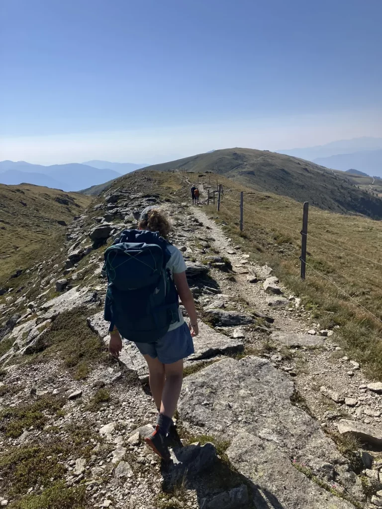 A hiker carrying a backpack walking along a rocky mountain ridge trail under a bright blue sky.