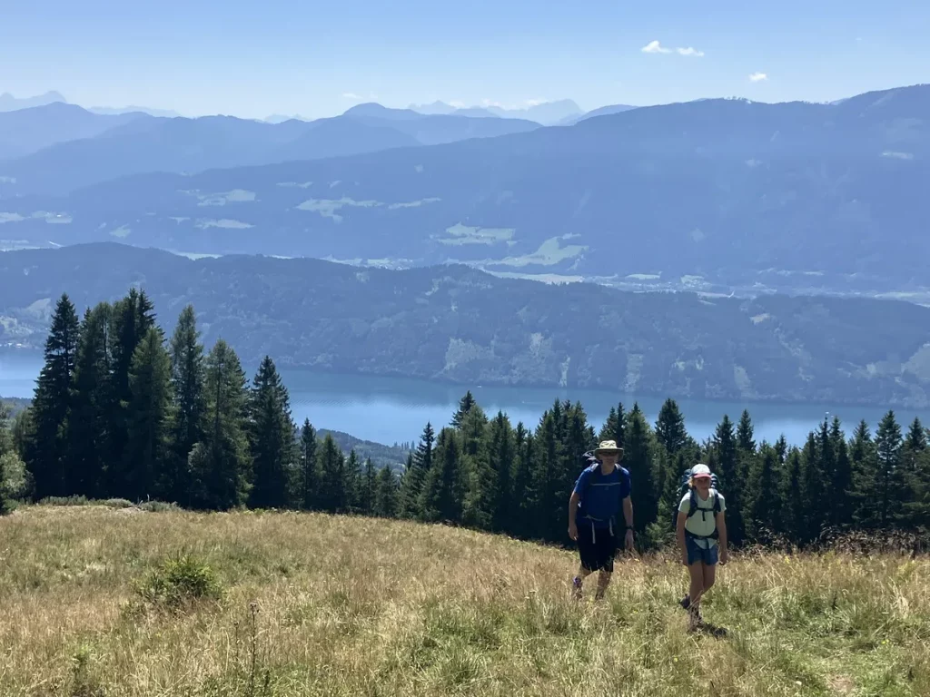 Two hikers walking uphill across a meadow with a lake and rolling mountain ranges stretching out below. A scenic moment from a beginner hut to hut hike in Austria.