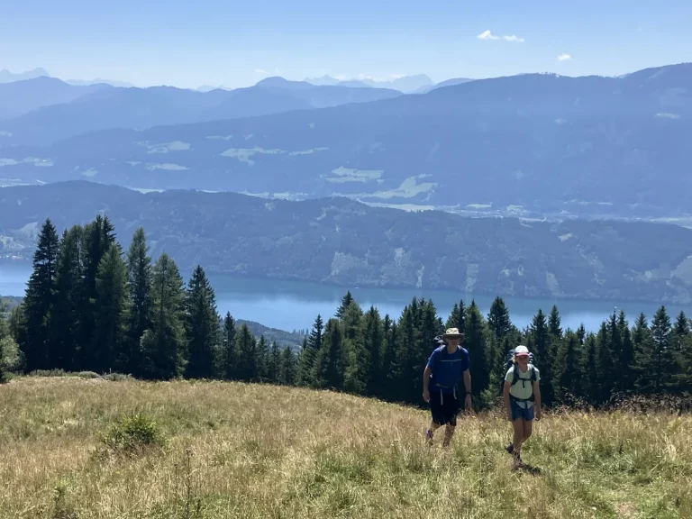 Two hikers walking uphill across a meadow with a lake and rolling mountain ranges stretching out below. A scenic moment from a beginner hut to hut hike in Austria.