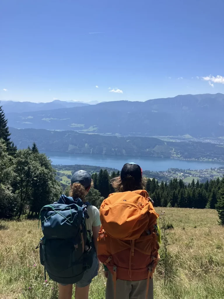 Two hikers with large backpacks standing on a grassy hillside looking out at a sparkling blue lake and mountain ranges in Austria. A quiet pause on a beginner hut to hut hike in Austria.