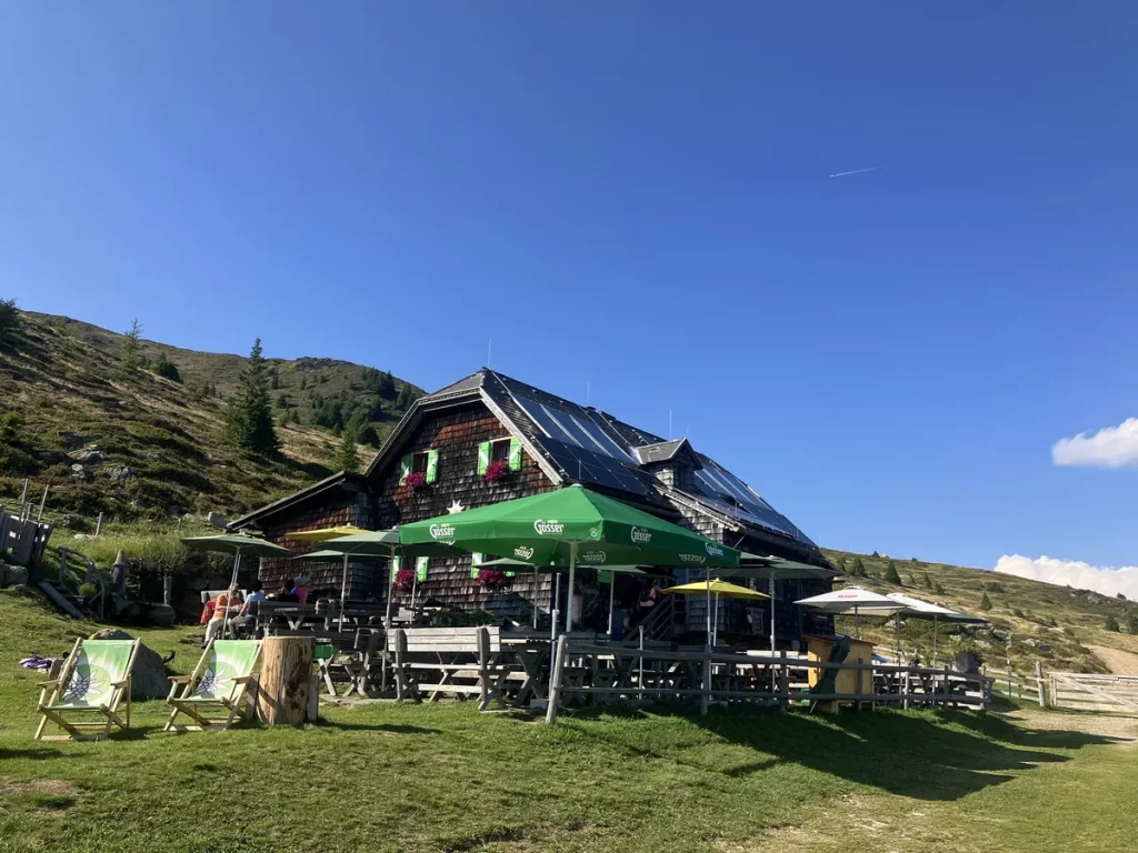 A rustic alpine hut with green umbrellas and flower boxes, set against rolling hills under a clear blue sky.