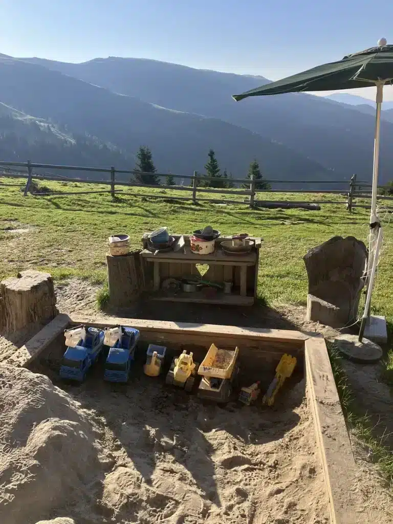 A children’s sandbox filled with toy trucks and diggers, plus a small mud kitchen, overlooking mountain scenery from a hut terrace.