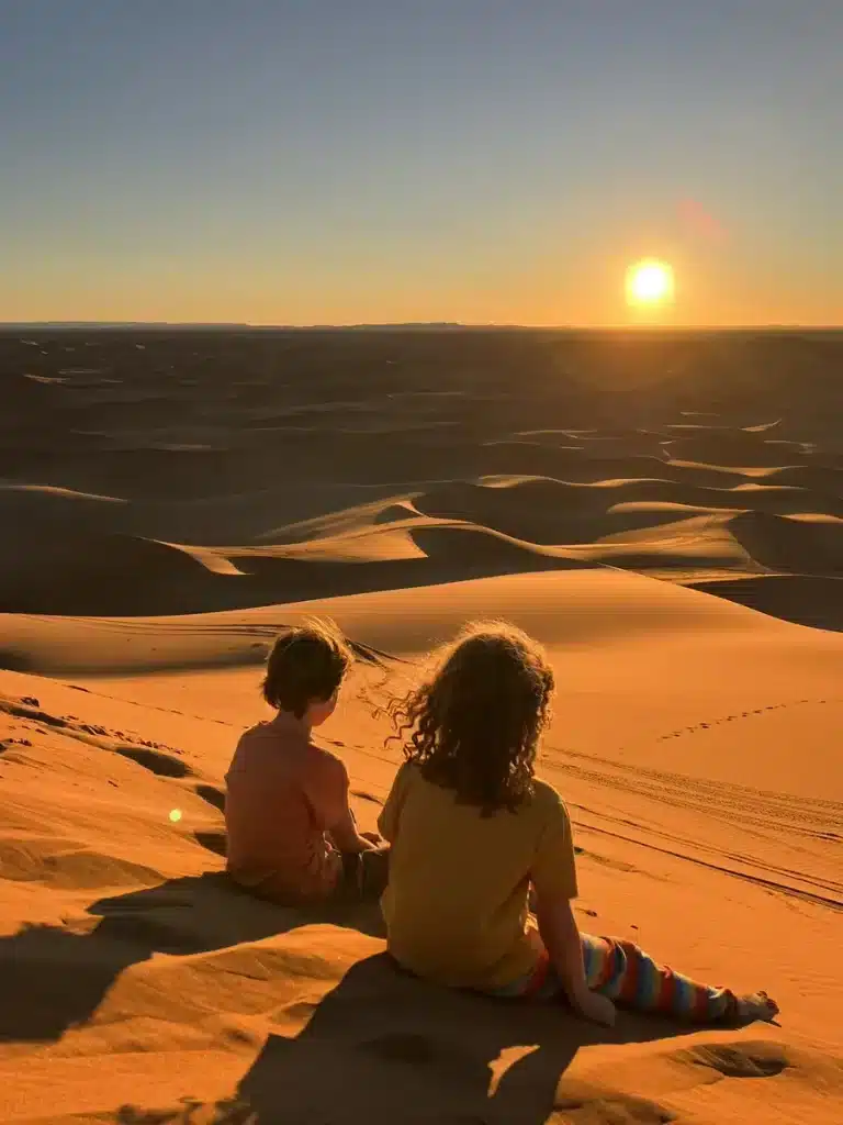 Two children sit side by side on a sand dune, watching the sun set over endless rolling sands in a peaceful desert landscape. Visiting Morocco is a magical way to unwind during October half term.