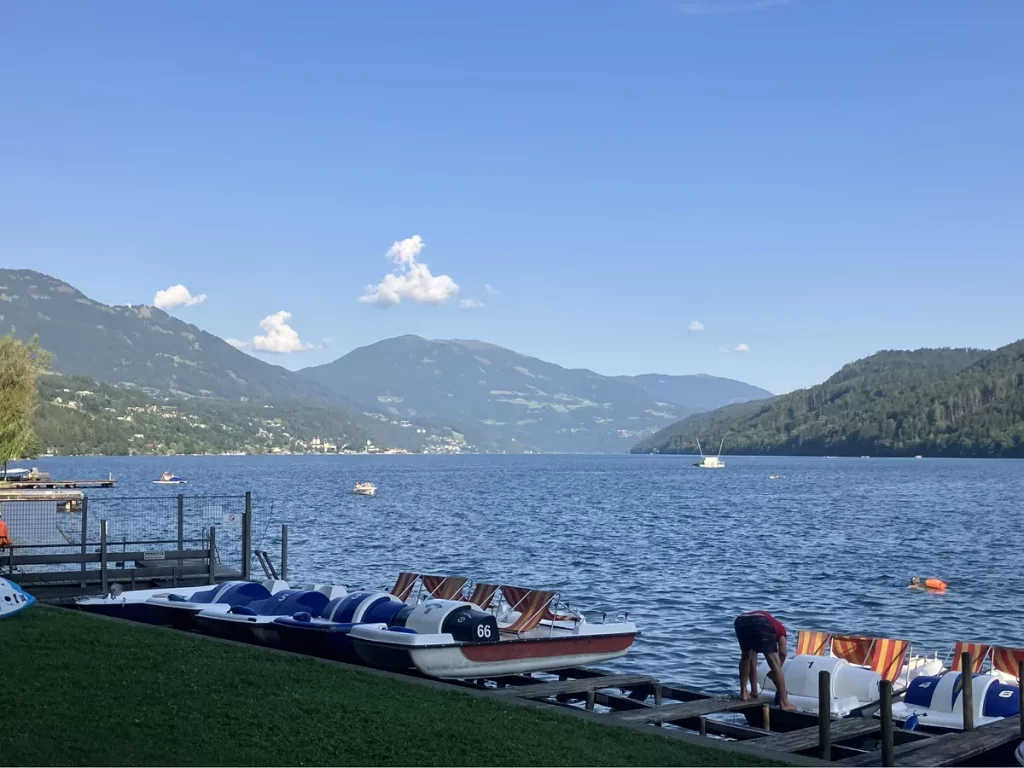 Pedal boats lined up at the edge of a lake (the Millstättersee) with mountains rising in the background and a few boats out on the water.
