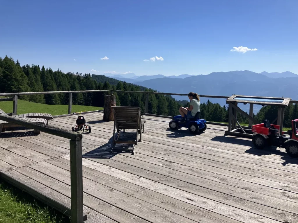 A child riding a toy tractor on a wooden deck with sweeping mountain views and forest below.