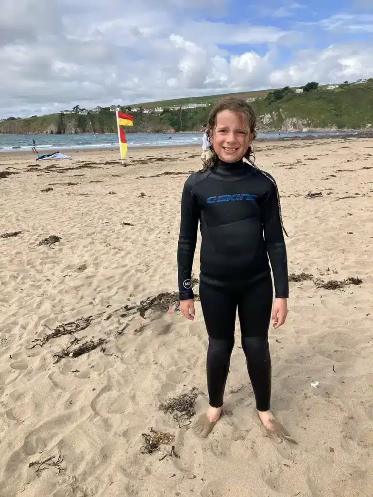 A girl in a wetsuit stands barefoot on a sandy beach, smiling after a surf session, with waves and coastal cliffs in the background. Beach adventures like surfing are great October half term ideas even in cooler weather.