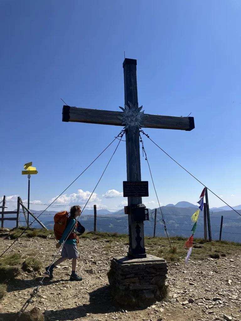 A hiker with a large orange backpack standing beside a summit cross decorated with prayer flags, looking out across the Austrian Alps.