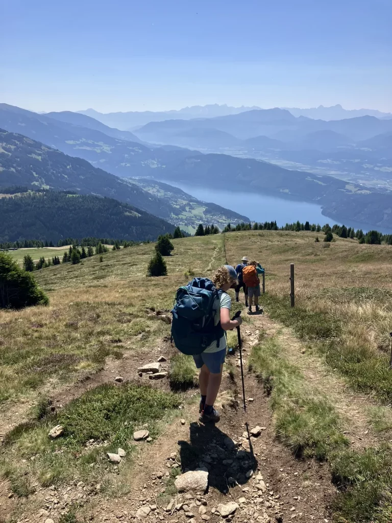 Hikers making their way down a grassy mountain trail with backpacks, overlooking a deep blue alpine lake and hazy ridges. A rewarding moment on a beginner hut to hut hike in Austria.