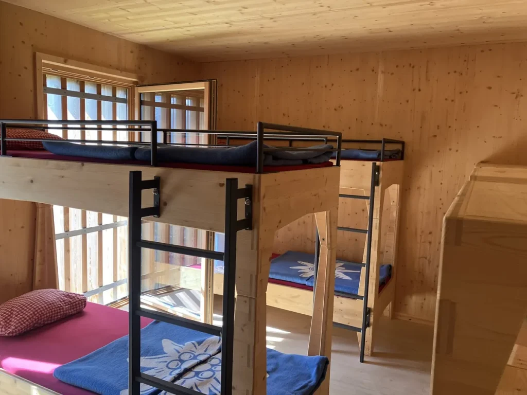 Inside a cozy alpine dorm room along the Wilder Kaiser hut to hut hike from Kufstein, four sturdy wooden bunk beds with blue edelweiss blankets and natural wood walls offer hikers a comfortable overnight stay.