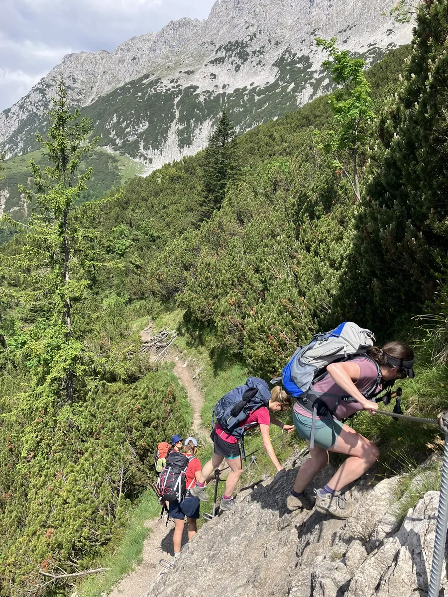Four women wearing large backpacks carefully climb a steep rocky section of trail using fixed metal cables for support. The exposed alpine terrain adds a challenging element to the group hike.