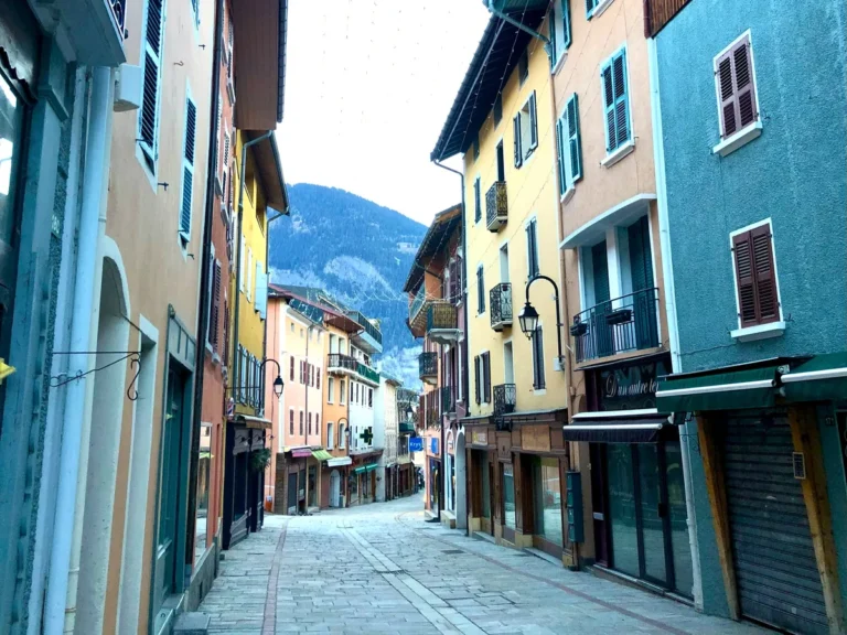 A quiet cobblestone street in Bourg Saint Maurice lined with colorful shuttered buildings and closed shops, with snow-dusted mountains visible in the background, capturing the peaceful charm of the town between days of Bourg Saint Maurice skiing.
