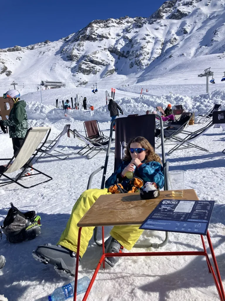 A child lounges in a sun chair with a drink in hand at a snowy outdoor café at Les Arcs, surrounded by mountains and ski gear under a bright blue sky. A ski menu and empty glasses sit on the snow-dusted table.