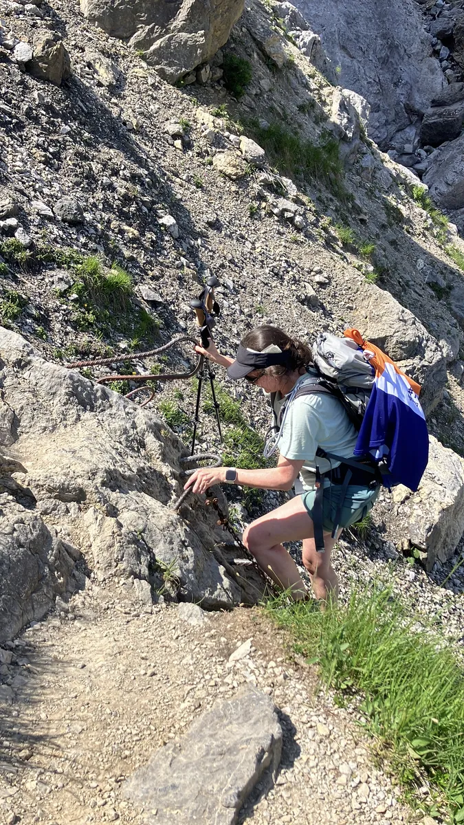 A female hiker climbs a rocky mountain path using secured metal cables and rungs, wearing a backpack and visor in bright sunlight.