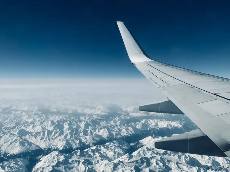 A view from an airplane window showing the aircraft’s wing against a deep blue sky. Below, a vast expanse of snow-covered mountain peaks stretches across the horizon, creating a dramatic and serene landscape.