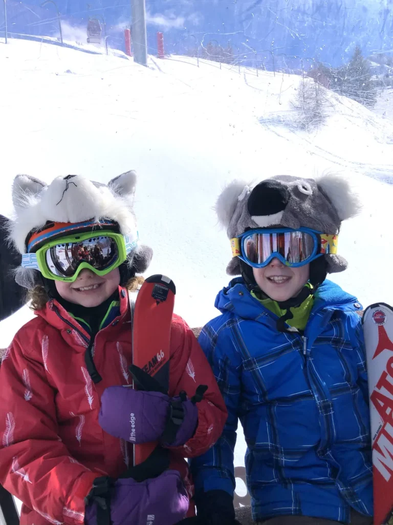 Two kids dressed for the slopes in colorful winter jackets, animal-themed ski helmets, goggles, and gloves, each holding their skis. Their layered ski gear highlights essential items to include on a family ski trip packing list.