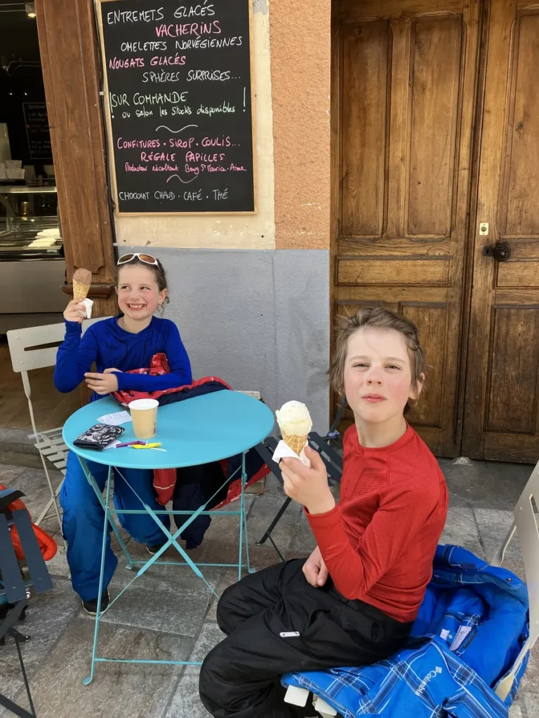 Two kids in ski clothing enjoy ice cream at a small blue café table outside a shop in Les Arcs. A chalkboard menu behind them lists desserts like omelettes norvégiennes and vacherins.