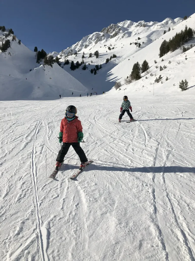 Two young children ski down a wide snowy slope between tree-lined mountain peaks at Les Arcs. The clear sky and shadows on the snow suggest perfect spring skiing conditions in April.