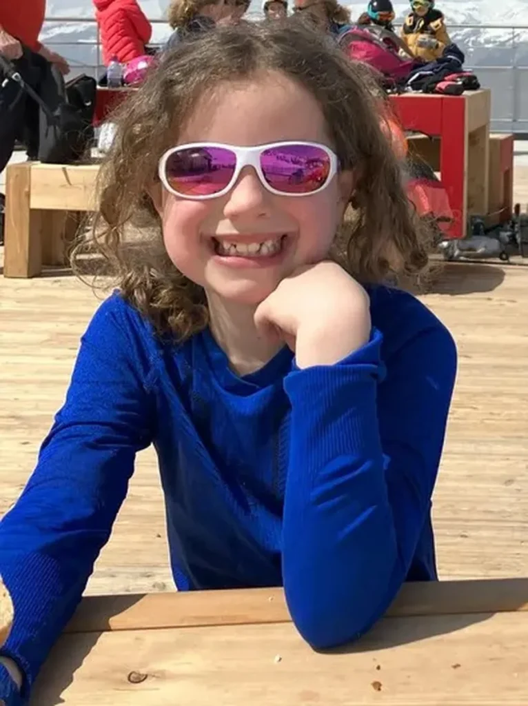 Smiling child in a bright blue thermal base layer top and white sunglasses, sitting at a wooden picnic bench in a ski resort area. Sunglasses and warm base layers are essentials to include on a ski trip packing list for sunny mountain days.