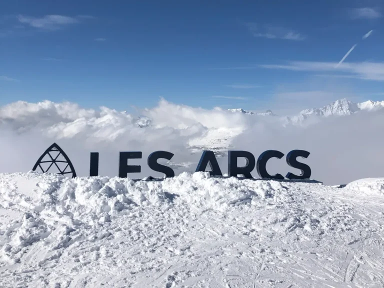 A large black “Les Arcs” sign stands atop a snowy ridge with clouds rolling over the mountains behind it. The scenic view marks a picture-perfect moment during a family ski holiday in Les Arcs.