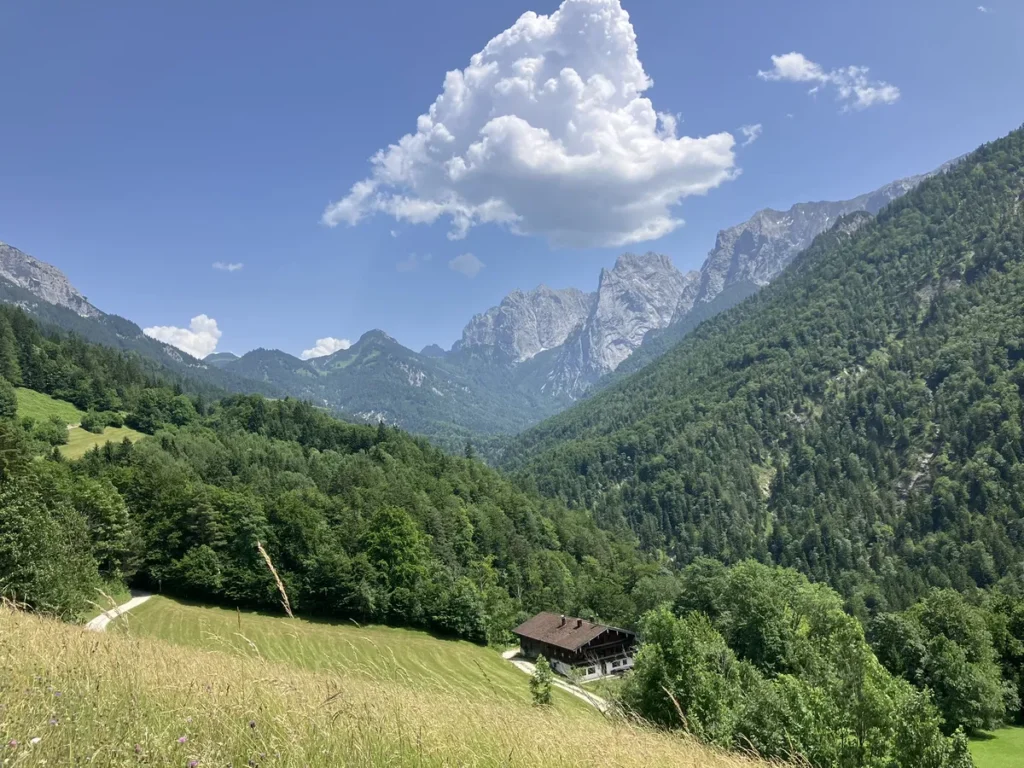 A quiet mountain valley with a wooden house nestled in a green meadow under towering jagged peaks, showcasing the tranquil beauty along the Wilder Kaiser hut to hut hike from Kufstein.