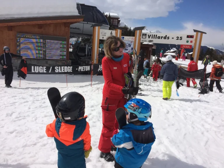 Ski instructor in red uniform talking to two young kids at the Villards ski lift entrance at Les Arcs Ski School, with skiers and signage around.