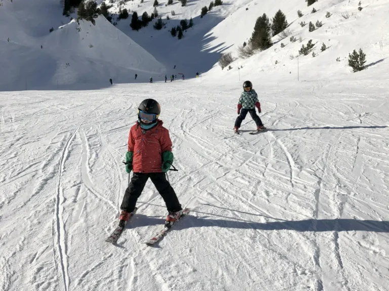 Two children in ski gear descend a wide, sunlit mountain slope under a clear blue sky. Their balanced postures show how kids can learn early, helping parents decide when children can start skiing.