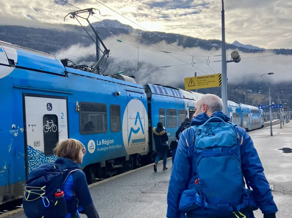 Travelers with backpacks board a blue “La Région Auvergne-Rhône-Alpes” train with snow-capped mountains in the background. Train access helps make affordable family ski holidays to Les Arcs convenient and eco-friendly.