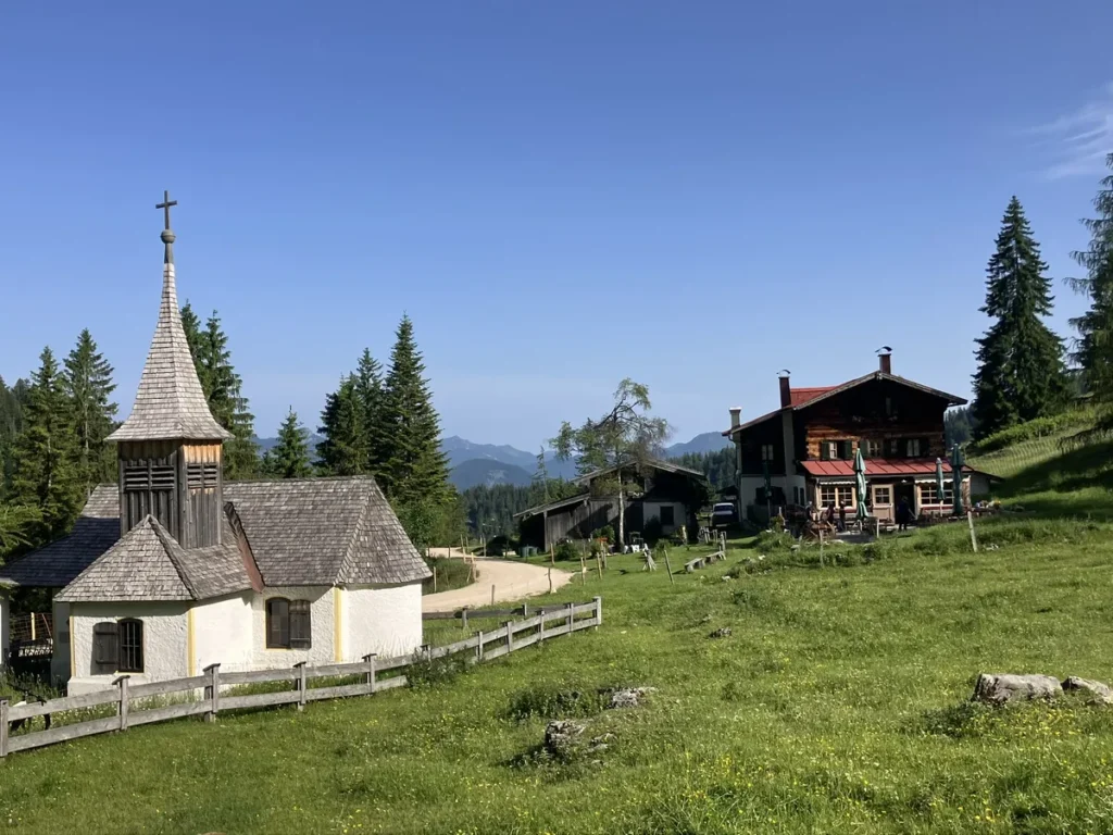 A small alpine chapel with a wooden steeple stands beside a traditional mountain guesthouse, surrounded by green meadows and pine trees along the Wilder Kaiser hut to hut hike from Kufstein.
