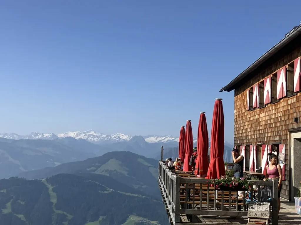 A wooden mountain hut with red-trimmed windows and bright red umbrellas sits dramatically on a balcony overlooking the Wilder Kaiser mountain range, a scenic rest stop on the Wilder Kaiser hut to hut hike from Kufstein.