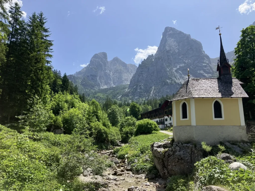 A picturesque alpine chapel and guesthouse sit side by side in a grassy clearing surrounded by pine trees, one of many charming scenes on the Wilder Kaiser hut to hut hike from Kufstein.