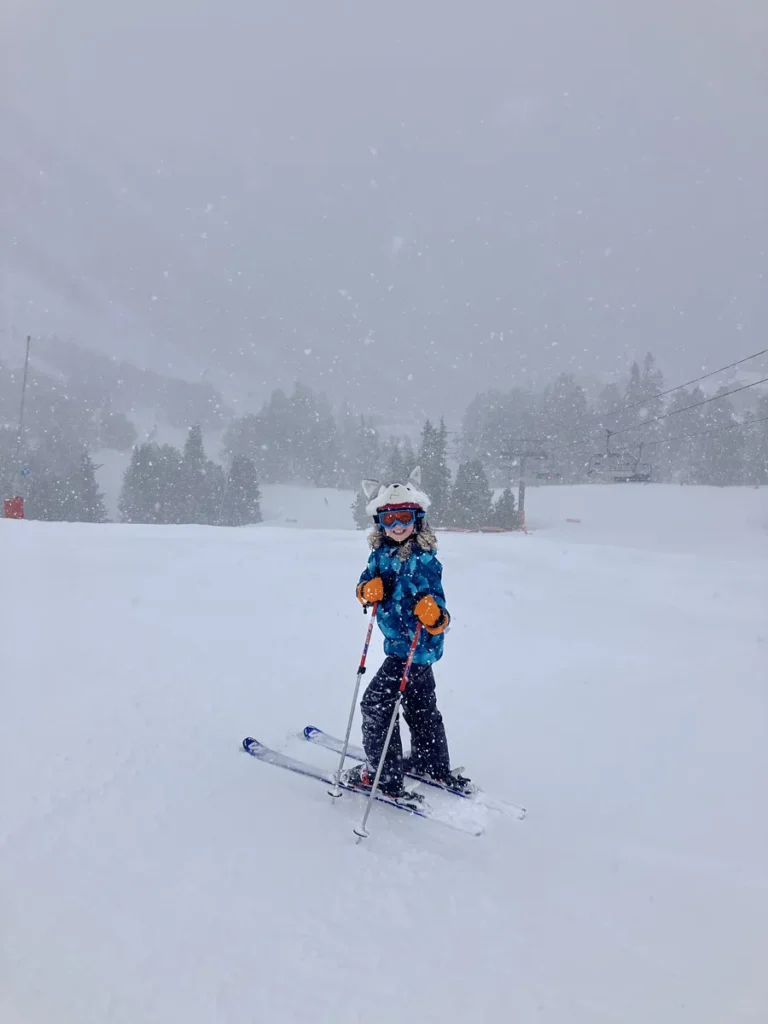 A child in bright ski gear stands on skis during a heavy snowfall, smiling while skiing at Les Arcs in April. Snow blankets the ground and trees, with visibility reduced by falling snowflakes.