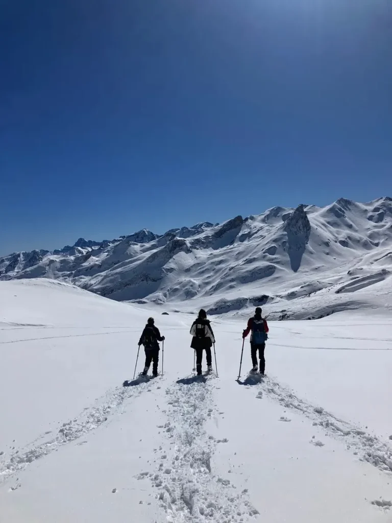 Three people snowshoeing through deep snow with trekking poles in the snowy mountains of Les Arcs under a clear blue sky. A scenic and active ski gift experience for outdoor lovers.