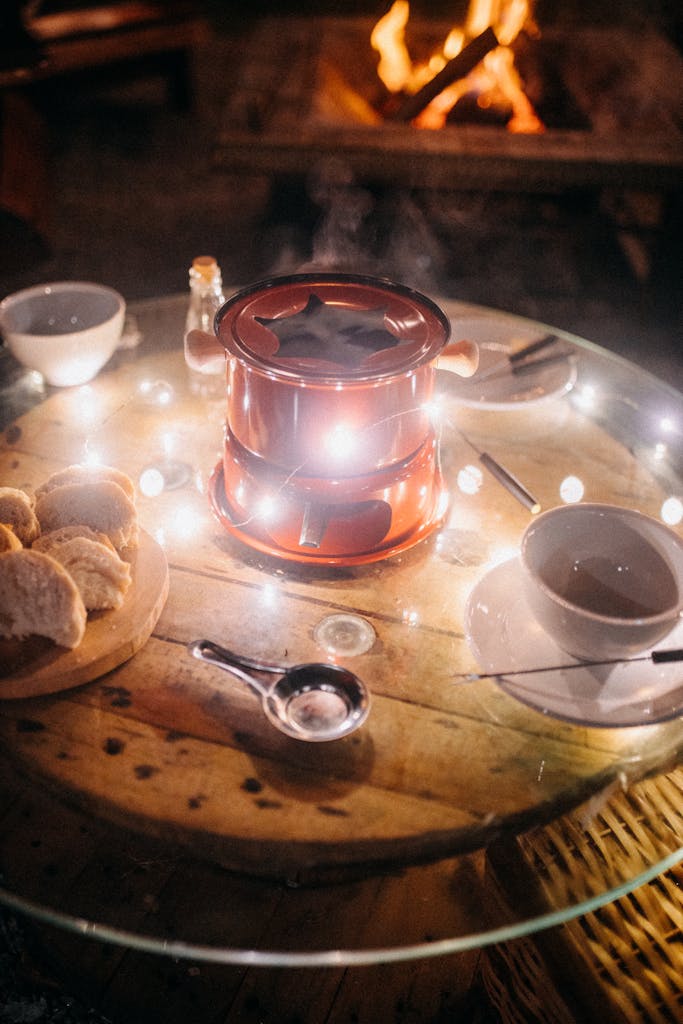 Warm fondue setup with bread on a rustic table, illuminated by string lights, outdoors by a fire.