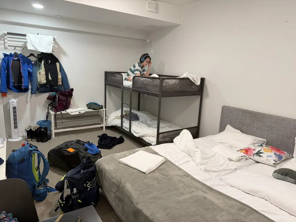 Simple hostel room with bunk beds, a double bed, backpacks on the floor, and jackets hanging on the wall. A child lounges on the top bunk, highlighting practical budget accommodations for a family trip.