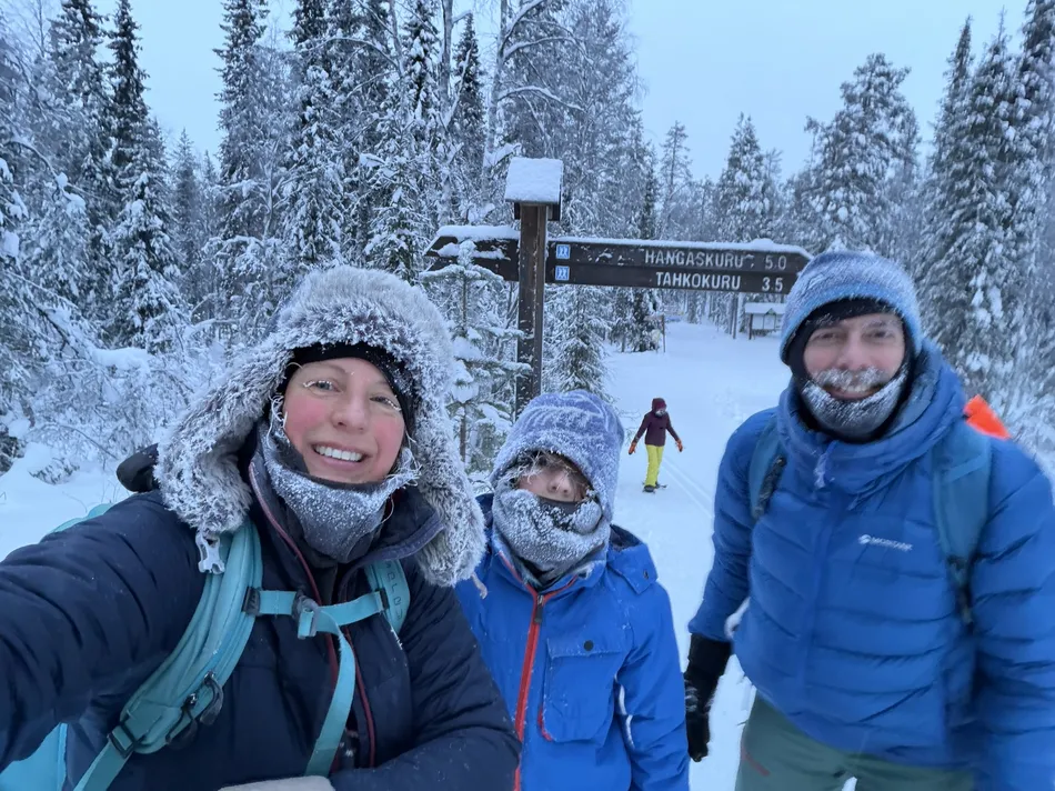 Family bundled in heavy winter clothing smiles for a snowy trail selfie, frost visible on hats and scarves. A wooden signpost stands behind them in a forest covered in deep snow.