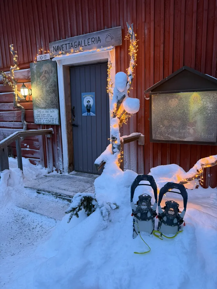 Snow covered red wooden building decorated with warm string lights and a sign reading Navettagalleria above the door. Snowshoes rest outside the entrance, suggesting a cozy winter stop during a cold weather adventure.