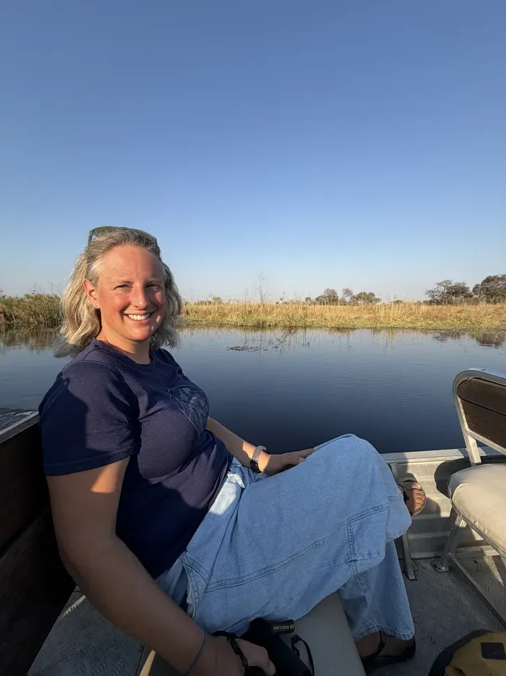 A woman relaxes on a small safari boat surrounded by calm water and tall golden reeds. She wears a short sleeve shirt and loose trousers for the warm sunny conditions.