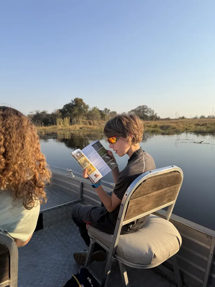 A child wearing sunglasses reads a wildlife guidebook while sitting on a safari boat. The field guide helps identify animals and birds during time on the water.