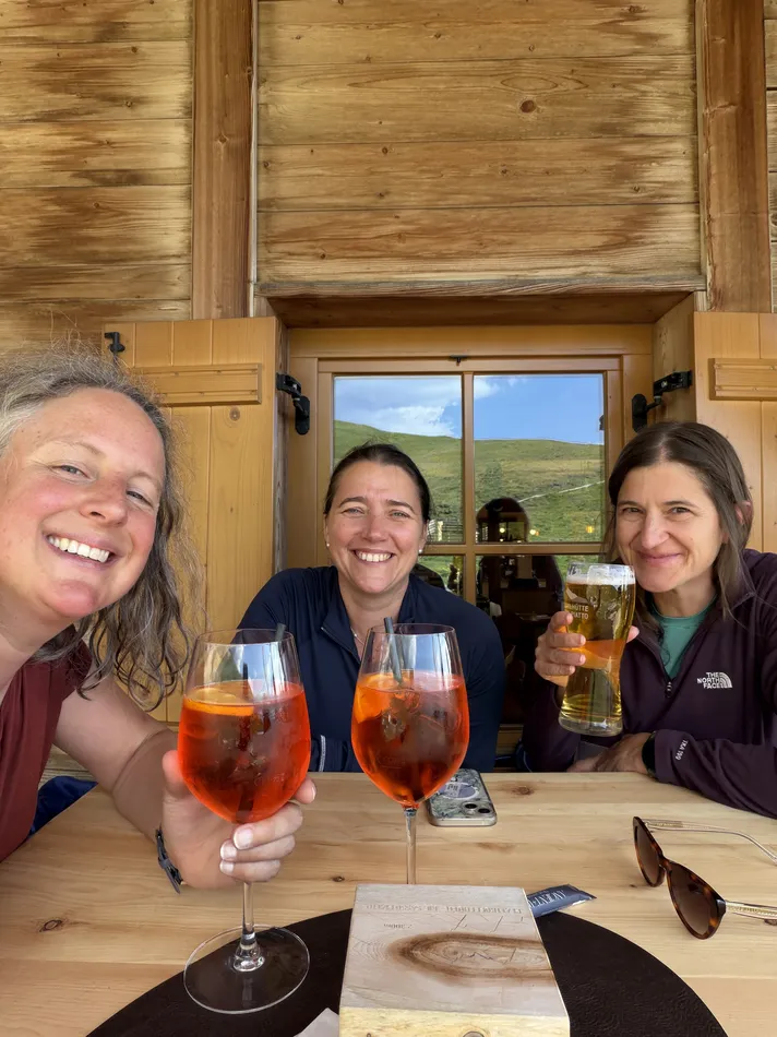 Three women smile together at a mountain hut table while holding bright orange Aperol spritz drinks and a beer. Relaxing together after a day outdoors is part of what makes hiking one of the best active girls trip ideas.