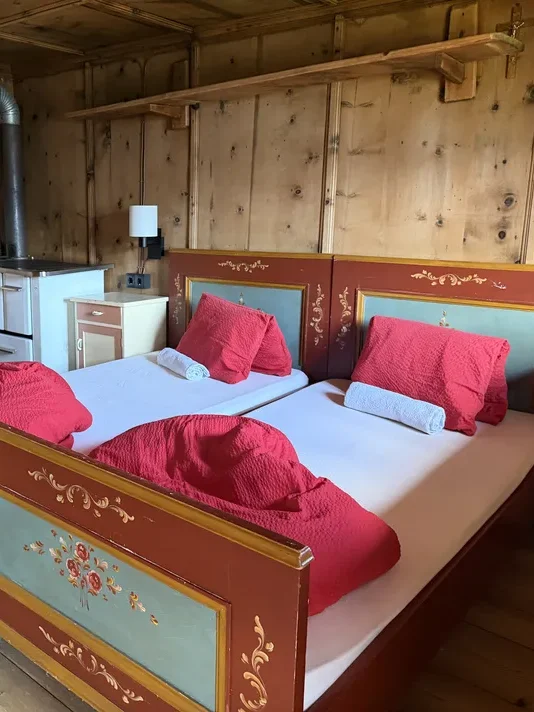 A rustic mountain hut bedroom has two painted wooden beds with red duvets and a small wood burning stove beside them. The simple timber room shows the traditional overnight accommodation available along the hiking route.